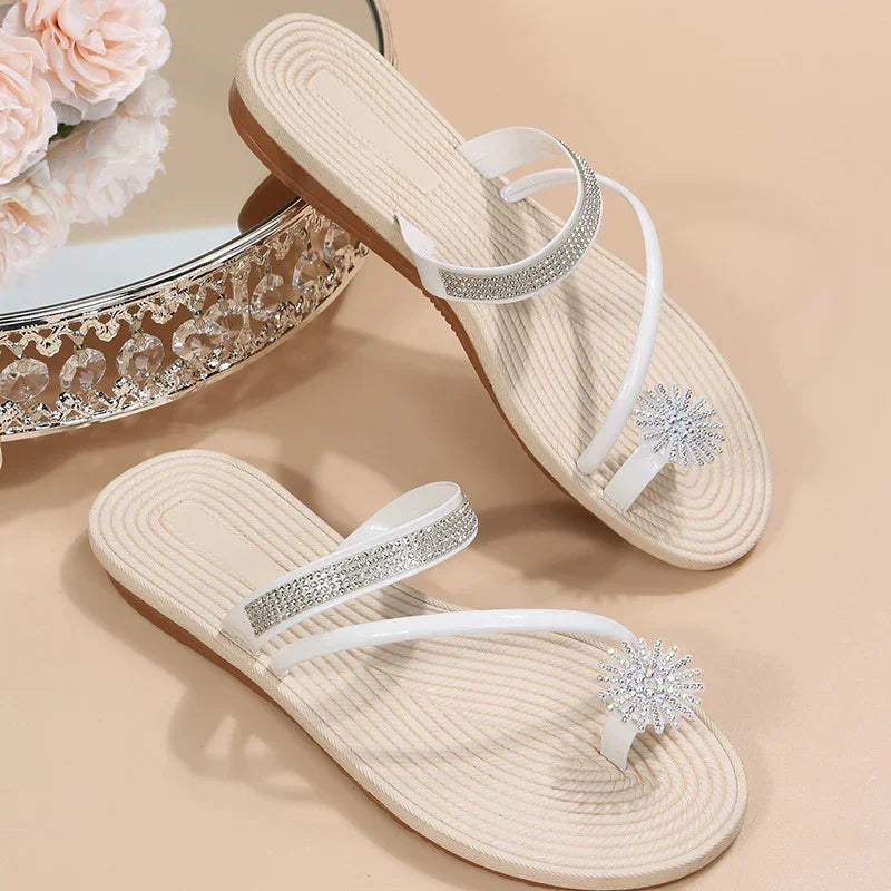 Elegant white flat sandals with rhinestone straps and floral jewel accents on beige background
