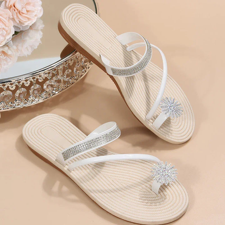 Elegant white flat sandals with rhinestone straps and floral jewel accents on beige background