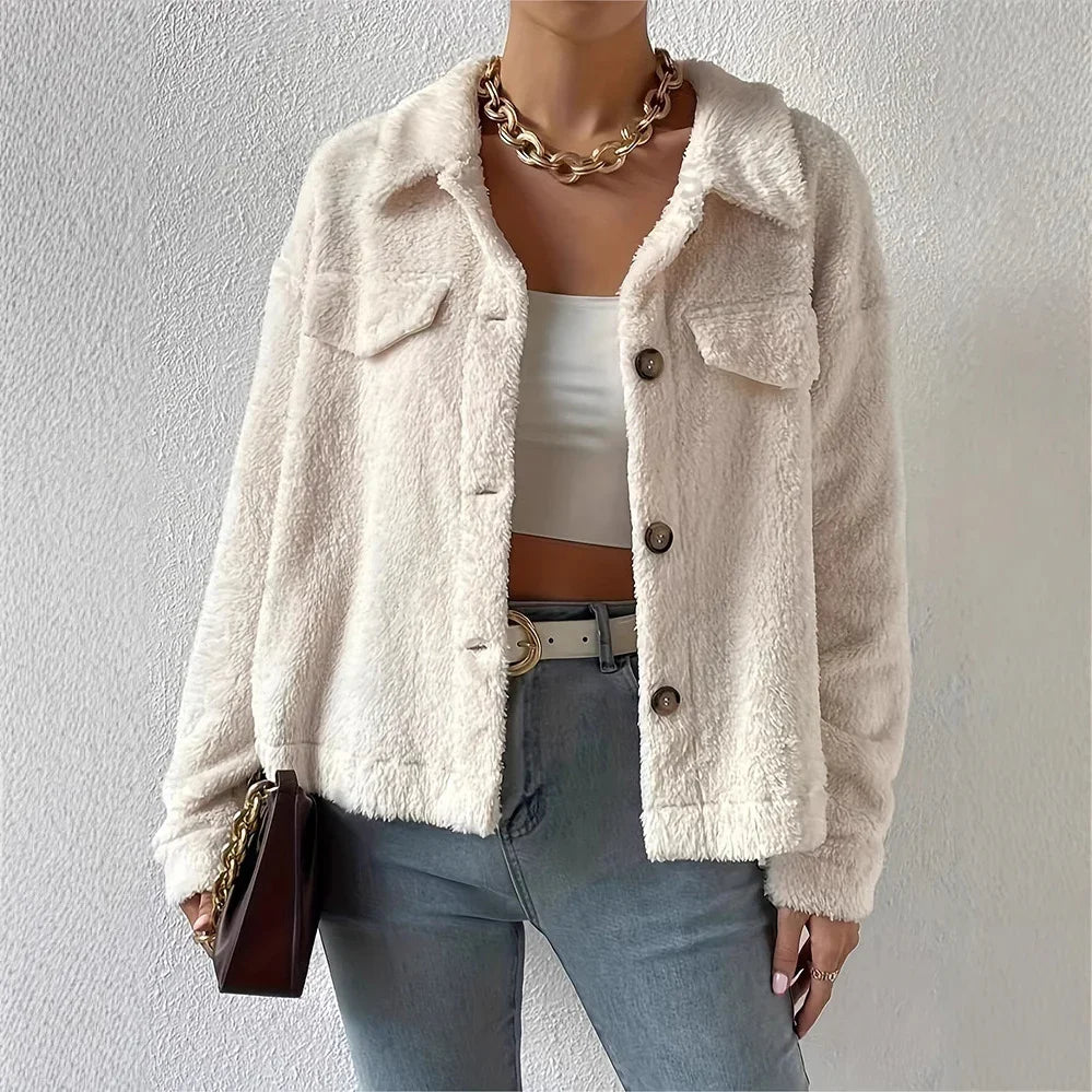 Woman wearing cream faux fur jacket, white crop top, light blue jeans, gold chain necklace, and holding brown bag
