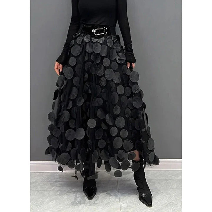 Woman wearing black maxi skirt with large circular appliques, black top, belt, and pointed black boots