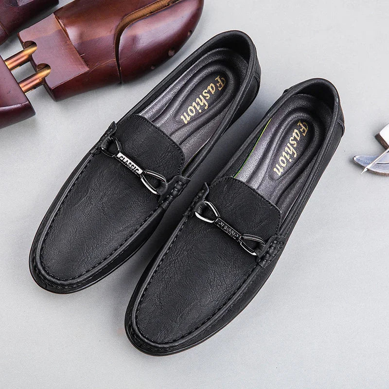 Black leather men’s loafers with metal buckle detail on gray surface beside wooden shoe trees