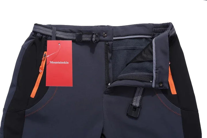 Dark grey outdoor hiking pants with black side panels, orange zipper pockets, adjustable belt, and Mountainskin tag