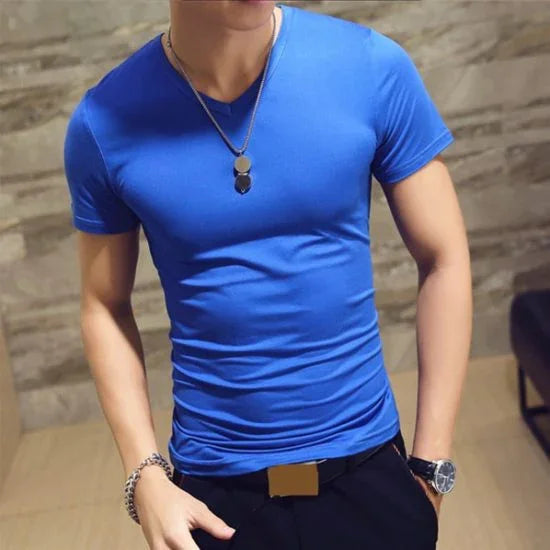 Man wearing fitted bright blue short-sleeve t-shirt, black pants with belt, silver necklace, bracelet, and watch indoors