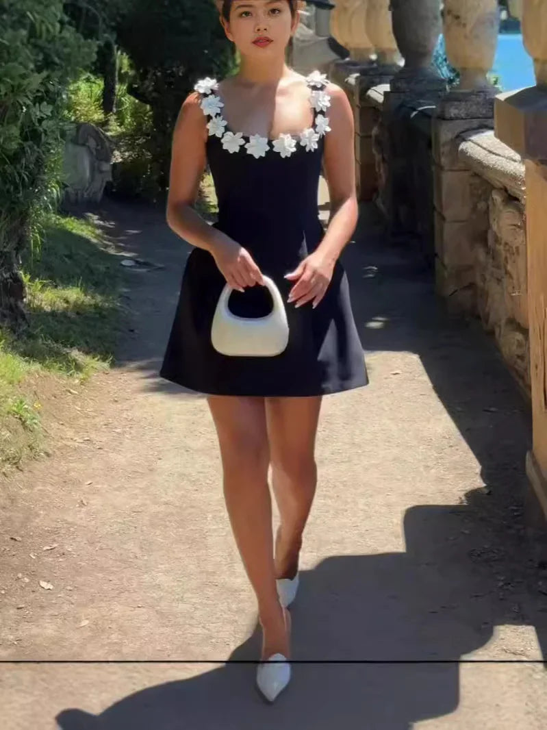 Woman wearing black dress with white floral straps holding white handbag walking outdoors on sunny day