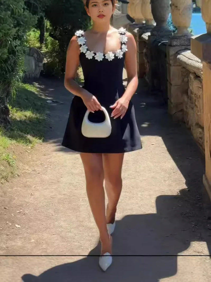 Woman wearing black dress with white floral straps holding white handbag walking outdoors on sunny day