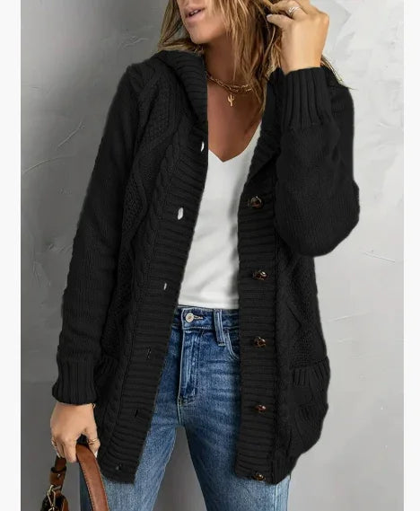 Woman wearing black cable knit cardigan with buttons, white top, and blue jeans against gray wall