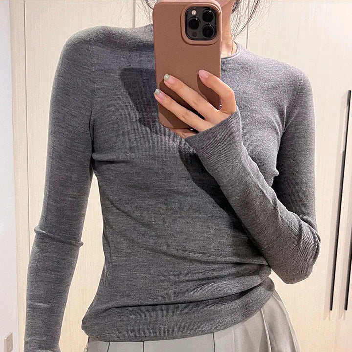 Person wearing fitted grey long-sleeve knit top taking a mirror selfie with smartphone indoors