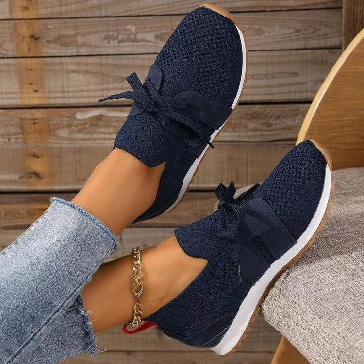 Navy blue knit sneakers with white soles and black laces worn with light blue jeans and a gold chain ankle bracelet