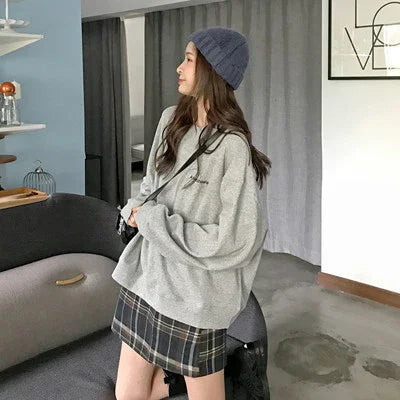 Woman wearing oversized grey sweater, plaid skirt, and blue knit hat standing in modern living room