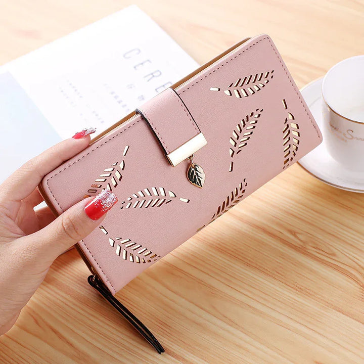 Pink leather wallet with leaf cutout design and gold leaf charm on wooden table with coffee cup