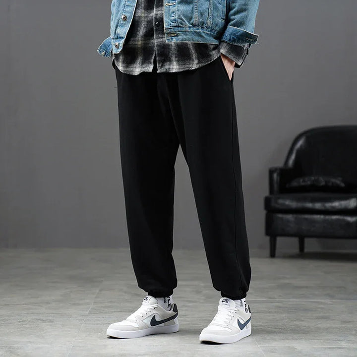 Man wearing black jogger pants, white sneakers, plaid shirt, and denim jacket indoors