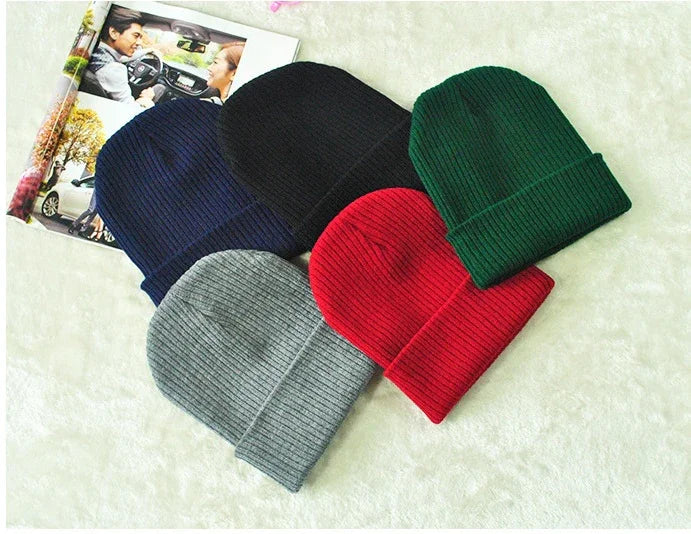 Five ribbed knit beanie hats in navy, black, green, gray, and red on a white surface with an open magazine nearby