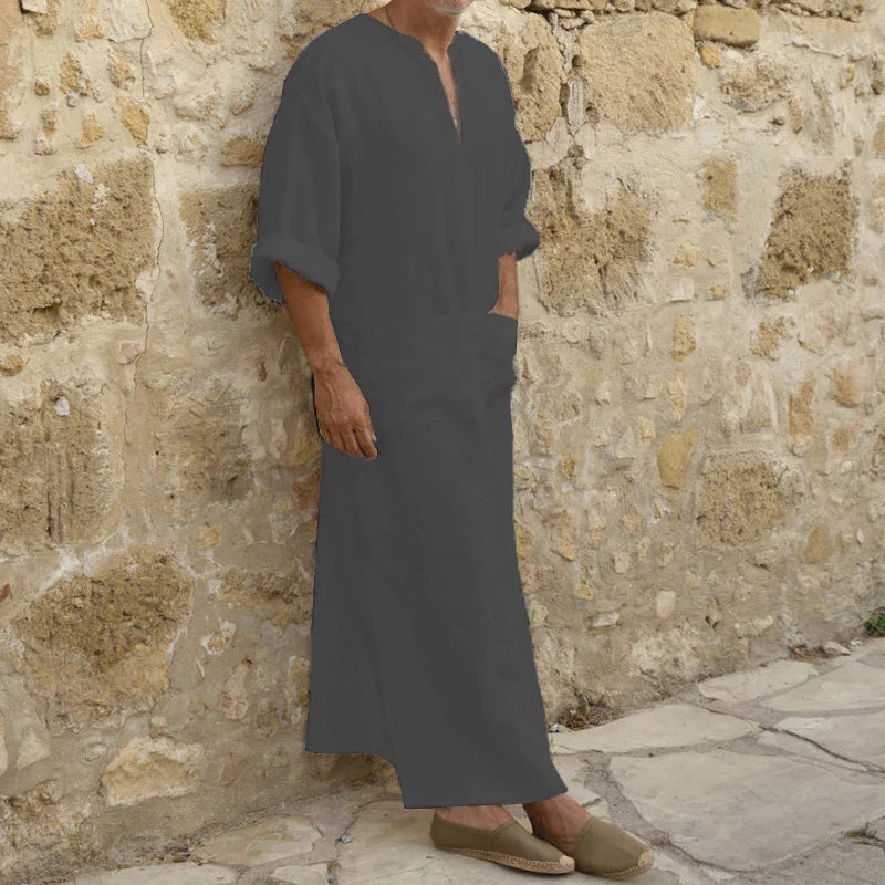 Man wearing dark gray traditional long robe and slip-on shoes standing against rustic stone wall