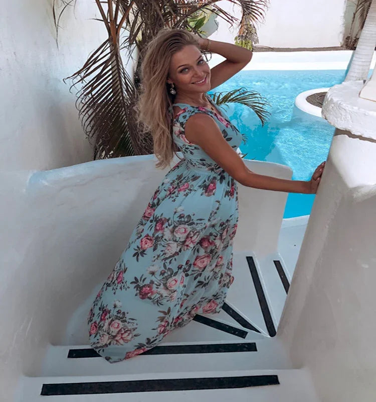Smiling woman in blue floral dress posing on white stairs near tropical pool and palm trees