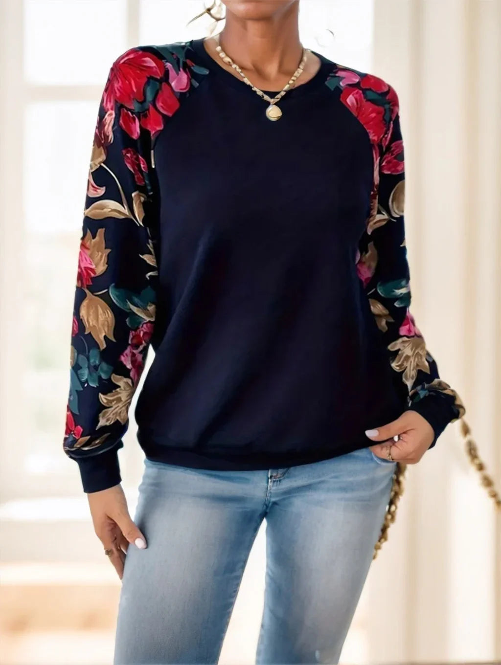 Woman wearing navy blue sweatshirt with vibrant floral print sleeves and light blue jeans indoors