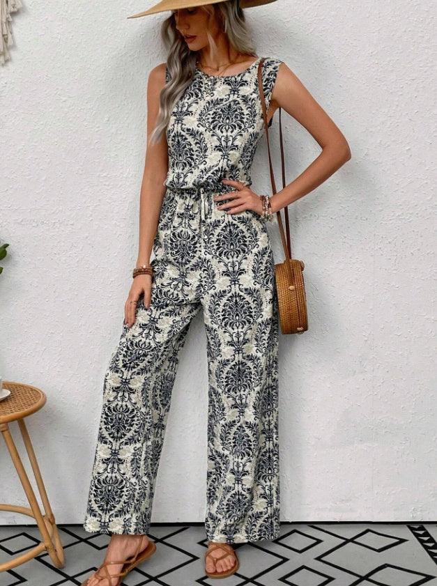 Woman wearing sleeveless black and white floral print jumpsuit with wide legs, straw hat, brown sandals, and wicker crossbody bag