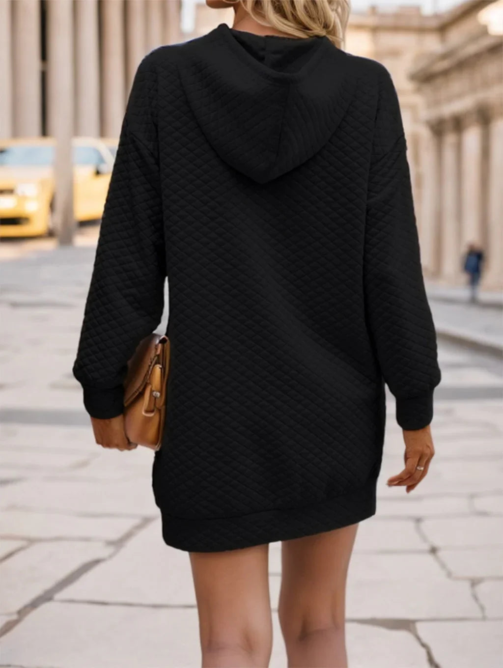 Woman wearing black quilted hoodie dress holding tan purse on city sidewalk