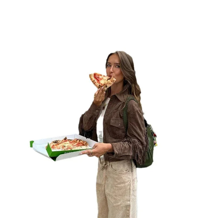 Young woman in brown jacket eating pizza slice from a green and white pizza box holding it with one hand
