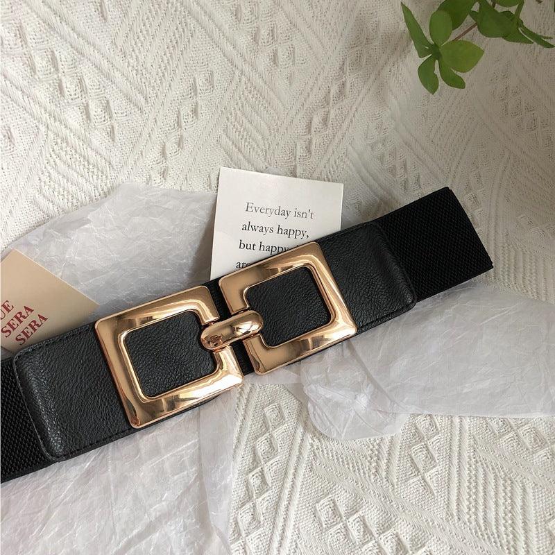 Black fashion belt with large gold rectangular buckle on textured white fabric background