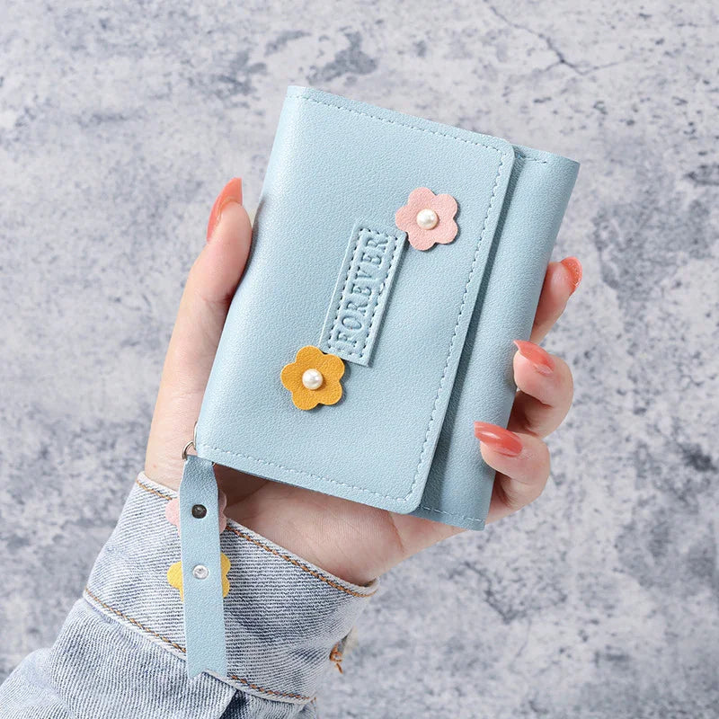Hand holding a light blue wallet with pink and yellow flower decorations and a wrist strap on a gray textured background