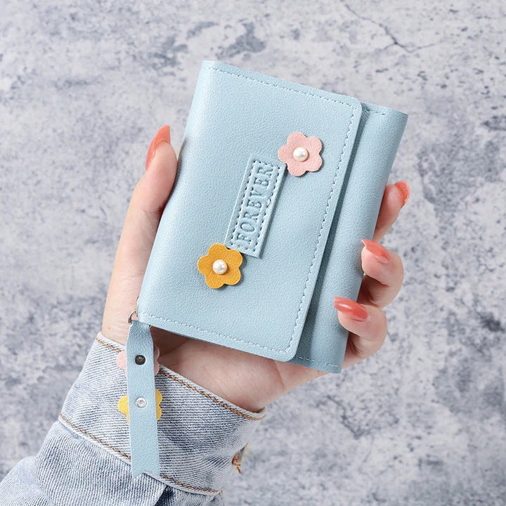 Hand holding a light blue wallet with pink and yellow flower decorations and a wrist strap on a gray textured background