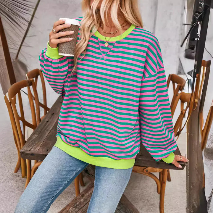 Woman wearing green and pink striped sweatshirt with jeans sitting on wooden bench holding coffee cup