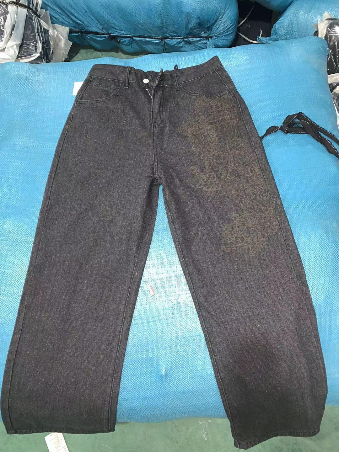 Black loose-fit denim jeans with faded text pattern on one leg, laid on blue fabric