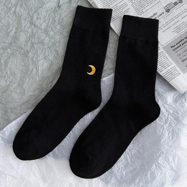 Black crew socks with an embroidered yellow crescent moon on one sock, displayed on crinkled fabric and newspaper background