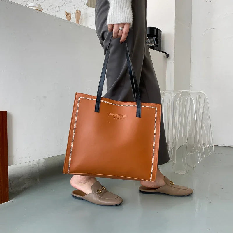 Person holding a large tan leather tote bag with black handles, wearing grey pants and beige slip-on loafers indoors