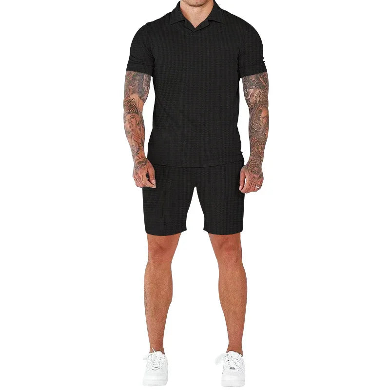 Man wearing black short-sleeve polo shirt and matching shorts with white sneakers, tattooed arms visible