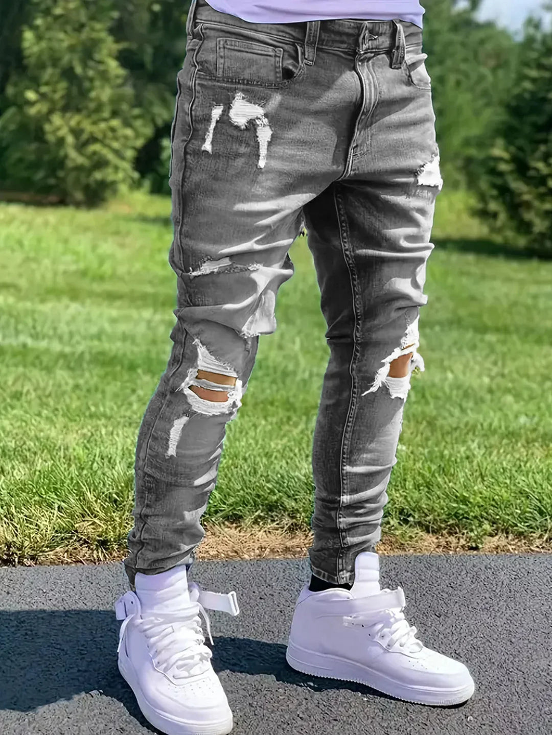 Person wearing ripped gray skinny jeans and white high-top sneakers standing on pavement with grass background