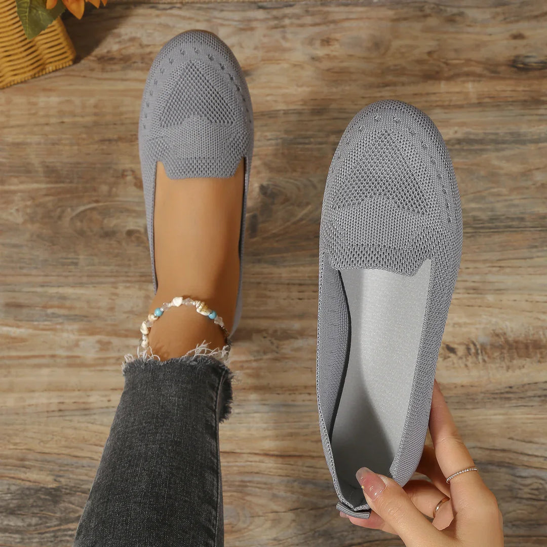 Light gray knitted ballet flats on wooden floor, one worn with an ankle bracelet and one held by hand