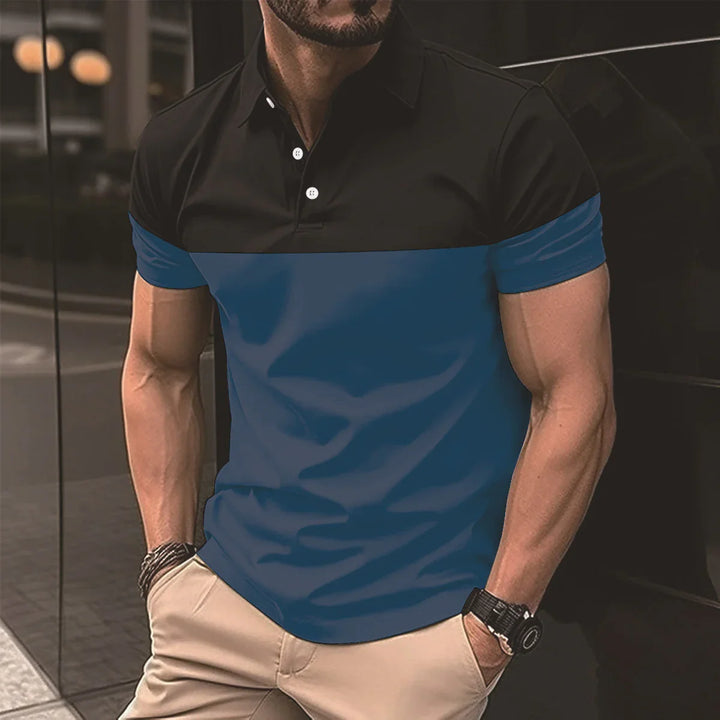 Man wearing black and blue color block polo shirt with beige pants standing against dark reflective wall