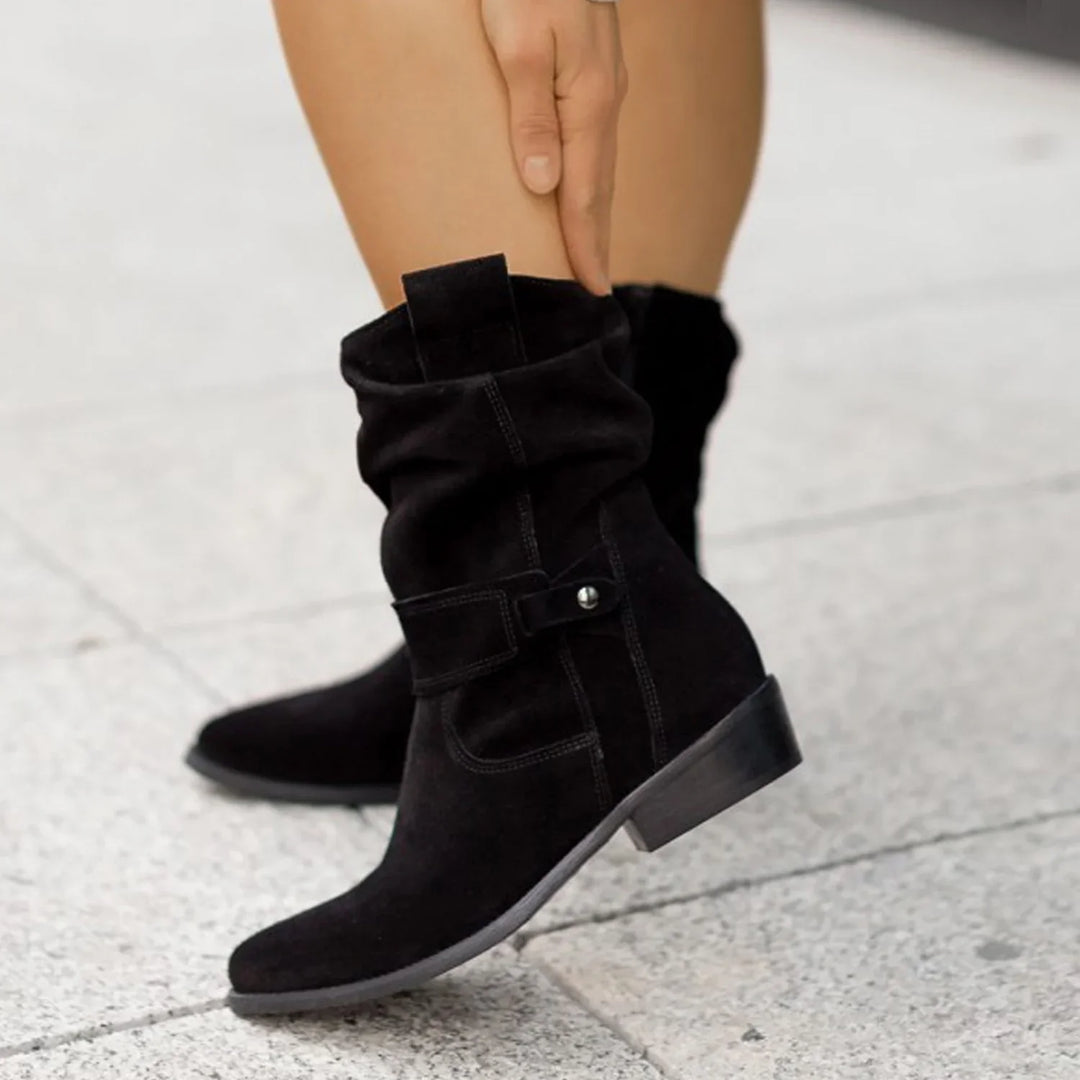 Black suede mid-calf boots with strap detail worn on pavement, hand adjusting boot top