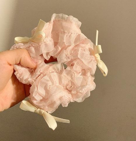 Hand holding a pink ruffled fabric scrunchie with cream-colored ribbon bows on a beige background