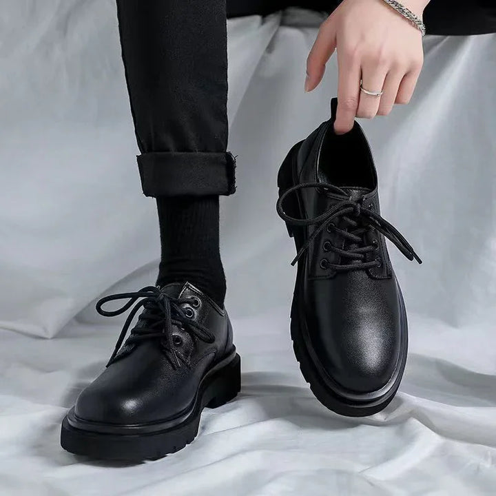 Pair of black leather lace-up shoes with thick soles, worn by person in black pants and socks