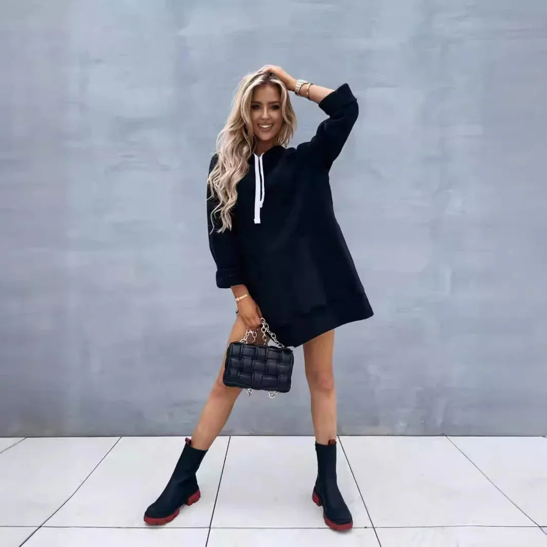 Blonde woman in black hoodie dress with white drawstrings, black boots, and quilted black handbag against gray wall