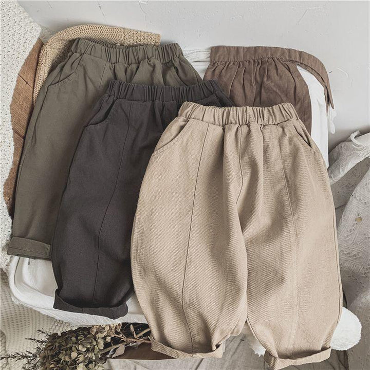 Four pairs of children's elastic waist pants in beige, dark gray, olive green, and brown laid out on a white surface with knitted textiles