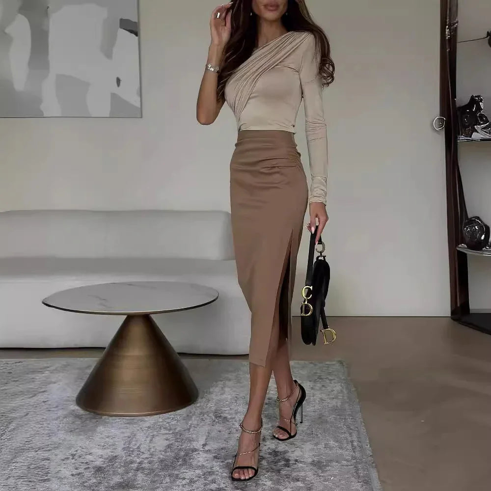 Woman in beige long-sleeve top and brown high-waist slit midi skirt holding black handbag indoors