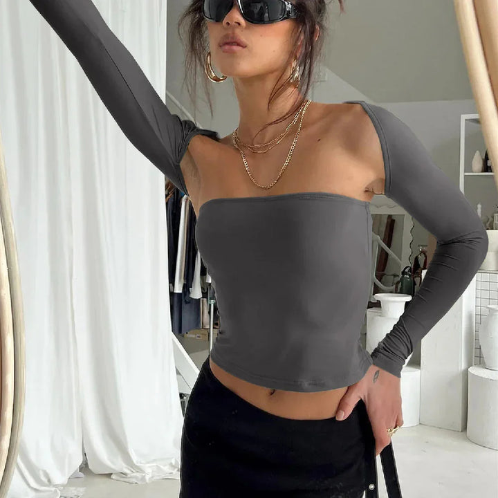 Woman in gray off-shoulder top and black skirt posing with sunglasses and gold jewelry in modern room