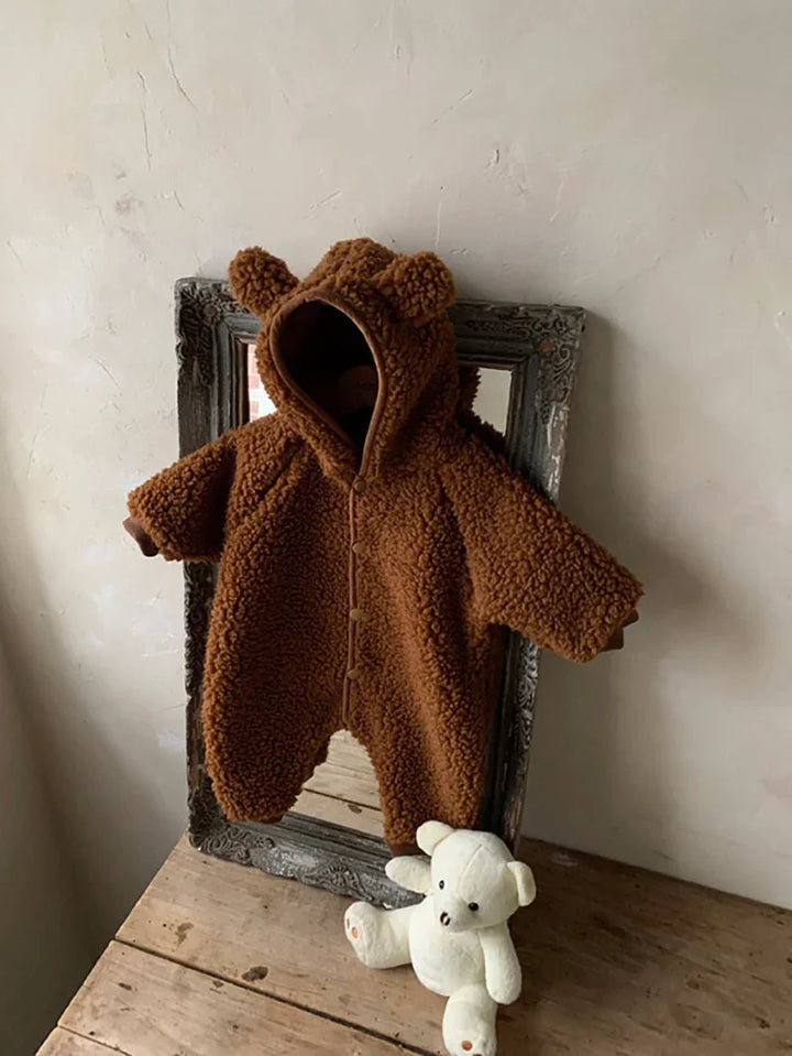 Brown teddy bear hooded baby jumpsuit hanging on ornate mirror with white teddy bear on wooden table