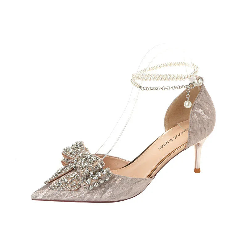 Elegant beige high-heeled shoe with pointed toe, rhinestone bow, and pearl ankle strap