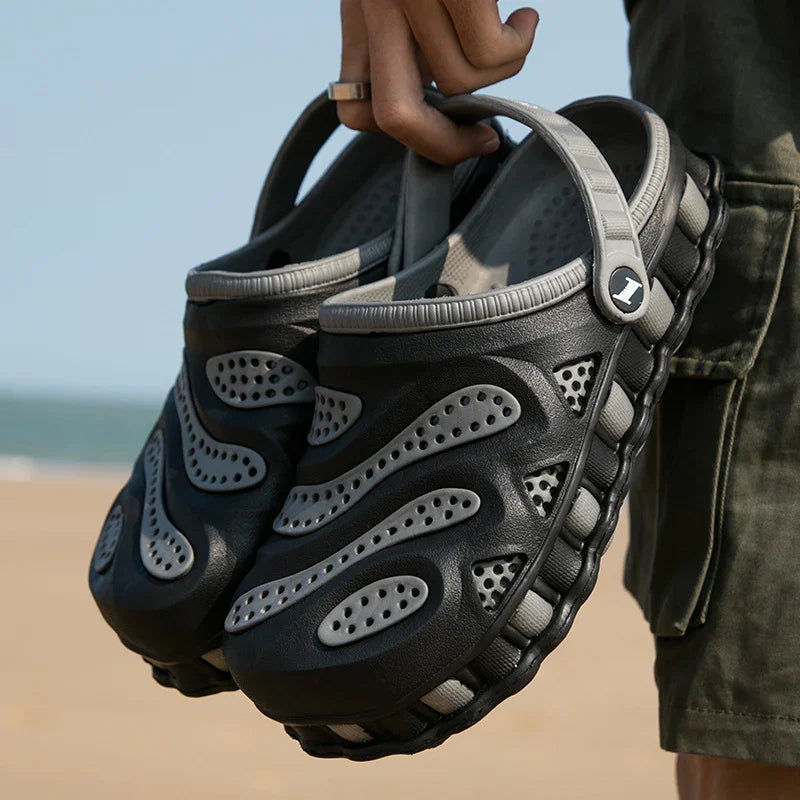 Person holding black and gray ventilated casual slip-on shoes with adjustable heel strap at beach