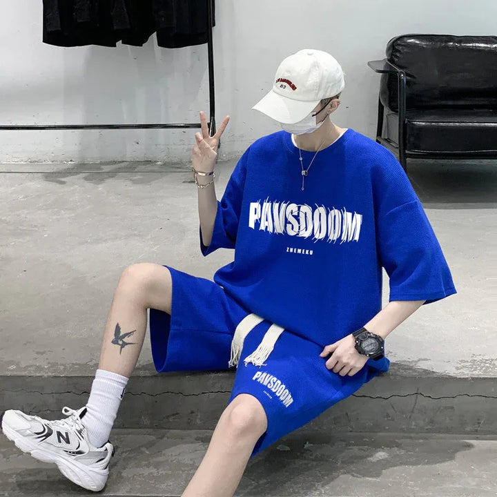 Person in blue PAWSDOOM outfit sitting on concrete ledge wearing white cap, sneakers, and showing peace sign