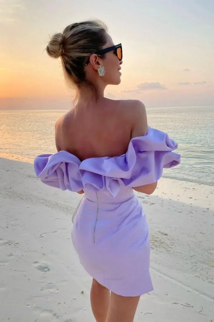 Woman in a lilac off-shoulder dress standing on beach at sunset wearing sunglasses and flower earrings