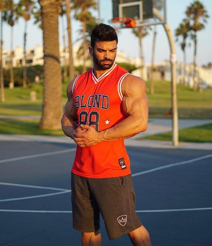 Muscular man in red sleeveless BLGND 88 jersey and gray shorts standing on outdoor basketball court with palm trees