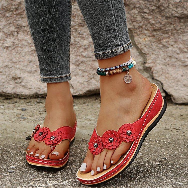 feet wearing red floral leather sandals and colorful beaded ankle bracelets on concrete ground