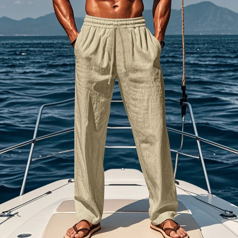 Man wearing beige linen pants and sandals standing on yacht deck with ocean and mountains background