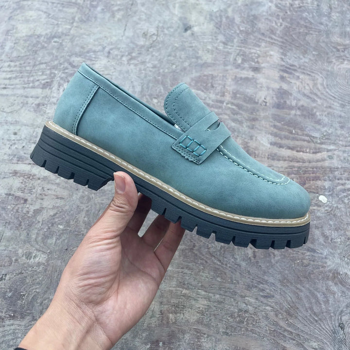 Hand holding a teal blue suede loafer with chunky black rubber sole on concrete floor