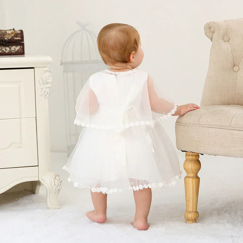 Baby girl in white lace dress standing barefoot, holding beige upholstered chair in light room